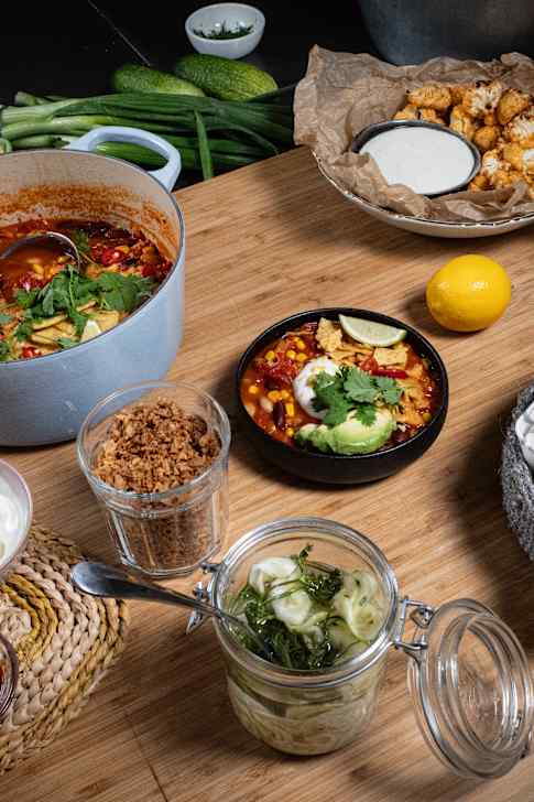 Puisella keittiötasolla on tortillakeittoa kattilassa ja lautasella, pieni kulho hapankermaa, paahdettua sipulia lasissa, lasipurkillinen säilöttyä kurkkua, kulhollinen paahdettuja kukkakaalin nuppuja, sitruuna ja leipäkori, jossa on kaksi täytettyä patongin puolikasta.