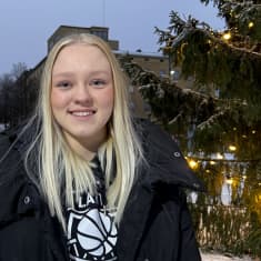 Vaaletukkainen nuori nainen katsoo suoraan kameraan ja hymyilee. Taustalla on iso joulukuusi.