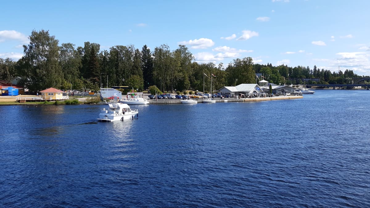 Vene menossa järveltä päin kuvattuna satamaan aurinkoisena kesäpäivänä. Satamassa autoja parkissa, liput liehuvat ja laiturissa on muita veneitä. Taustalla näkyy silta.