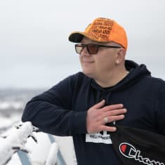 Oranssiin lippikseen ja siniseen huppariin pukeutunut mies nojaa korkealla sijaitsevaan kaiteeseen ja katselee kaukaisuuteen. Taustalla näkyy kaupunkimaisemaa.
