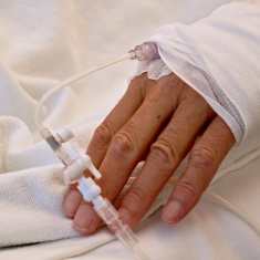 Photo shows the hand of a patient as they lie in a hospital bed.