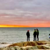 Tre personer står på klipporna vid havet i solnedgången.
