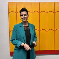 Riia Martinoja katsoo kameraan. Hänen takanaan on oranssi-punainen maalaus.