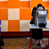 Ihmisiä äänestyskoneiden takana värikkään kaapiston edessä