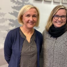 Lähikuvassa kaksi vaaleahkohiuksista naista, vasemmalla polkkatukkainen nainen sinisessä villatakissa ja oikealla hiukan nuorempi silmälasipäinen ja pitkätukkainen nainen. Molemmat hymyilevät. 