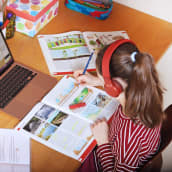 En flicka sitter vid ett skrivbord och skriver i en skolbok. Framför henne en dator.