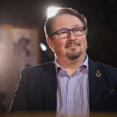 Profile of with short brown hair and goatee beard, wearing glasses and a dark suit jacket with a checkered shirt.