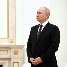 Putin seisoo kuvan oikeassa laidassa kädet ristissä edessään odottavan näköisenä. Hänellä on musta puku ja harmaa raidallinen kravatti. Huoneen seinät ovat vaaleat. Takkarakennelman päällä on koristeellinen iso kello.