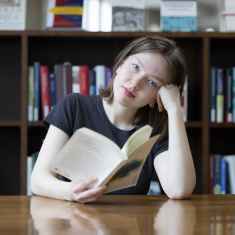 Ung kvinna tittar in i kameran med en uppslagen bok framför sig i biblioteksmiljö