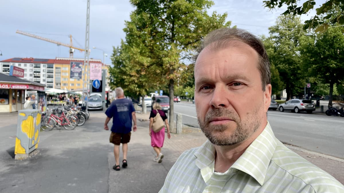 Mies vaaleassa kauluspaidassa katsoo kameraan vakavana. Taustalla näkyy Tampereen Tammelaa, ja kadulla käveleviä ihmisiä.