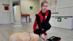 Punaiseen liiviin pukeutunut SPR:n työntekijä Eveliina esittelee sydäniskurin käyttöä. Lattialla ensiapunukke, sydäniskuri ja kyykyssä oleva Eveliina, joka painamassa iskurin nappia.