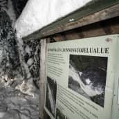 Hepokönkään luonnonsuojelualueen infotaulu lumisessa maisemassa ja taustalla hirsinen käymälä.