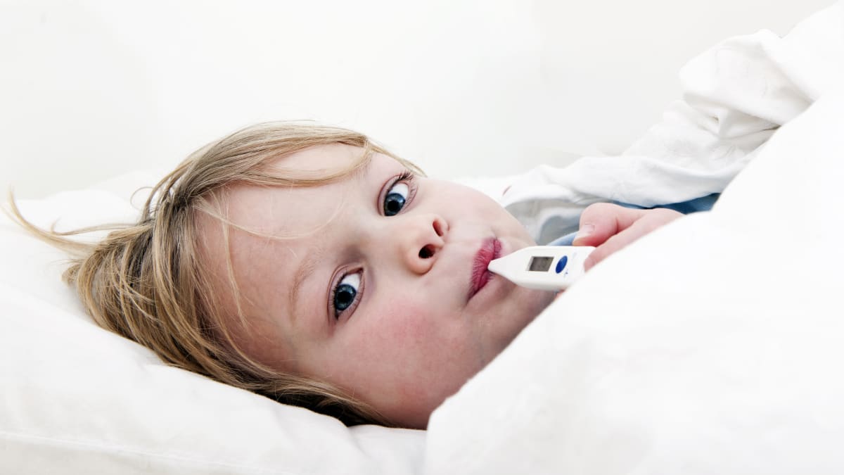 Ett barn ligger till sängs med feberröda kinder och en febertermometer i munnen.