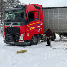 Punainen Viroon rekisteröity rekkaveturi poikittain lumisella tiellä, keula osittain pientareen puolella, kuljettaja levittää auton vieressä lumiketjuja.