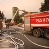 Paloautoja betoniaseman pihalla. Pihalla palomiehiä, jotka katsovat, kun toiset palomiehet ovat palovaurioita kärsineellä katolla.