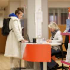 A woman in a white coat and carrying a backpack standing at a counter in an office reception area where another woman is seated.  A shelf of brochures is seen in the foreground.
