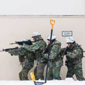 Neljä maastopukuihin ja kypäriin pukeutunutta ihmistä rynnäkkökiväärien kanssa menossa oikealta vasemmalle.