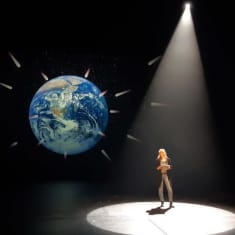 Nainen seisoo näyttämöllä, taustakankaalle heijastettu Maapallo.