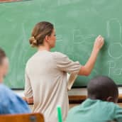 I förgrunden elever som sitter vid pulpeter, i bakgrunden en lärare som skriver på en tavla.