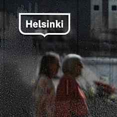 A window with the logo of the City of Helsinki. People walking by on the street are in the window's reflection.