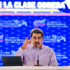 Maduro puhuu ja vahvistaa sanojaan kohottamalla oikeaa kättään. Hänellä on yllään vaalea kauluspaita. Hänen edessään pöydällä on mikrofoni ja näyttö. Taustakangas on sininen.