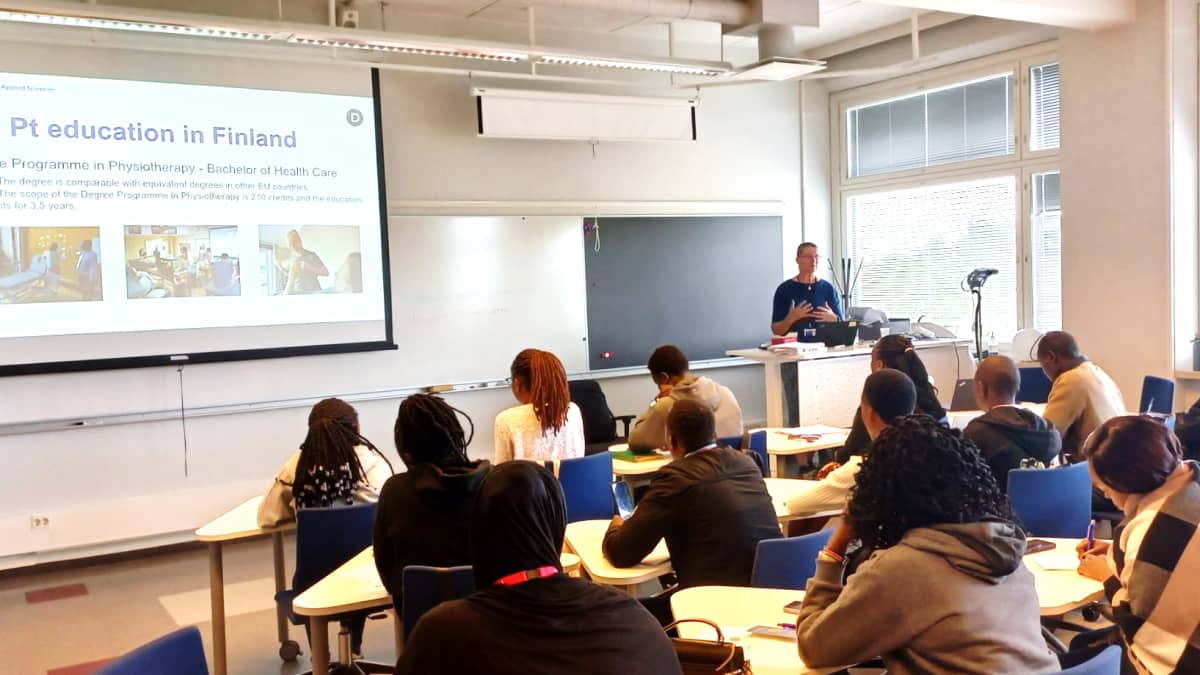 Kenialaisia opiskelijoita suomalaisessa luokkahuoneessa, opettaja puhuu luokan edessä. 