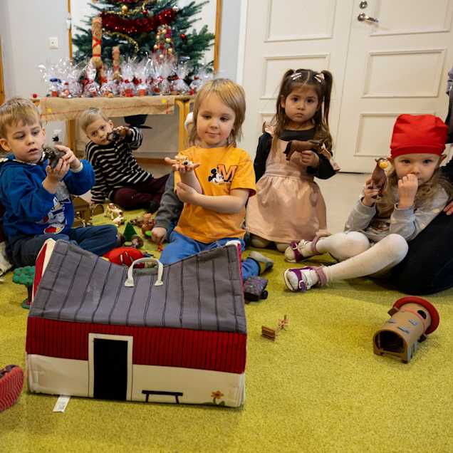 Lapset istuvat ympyrässä ja leikkivät pienillä taloilla ja eläimillä. Taustalla näkyy koristeltu joulukuusi.