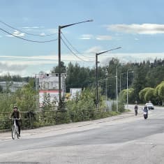 Tiellä kulkee muutamia autoja ja mopo, tien reunassa muutama pyöräilijä.