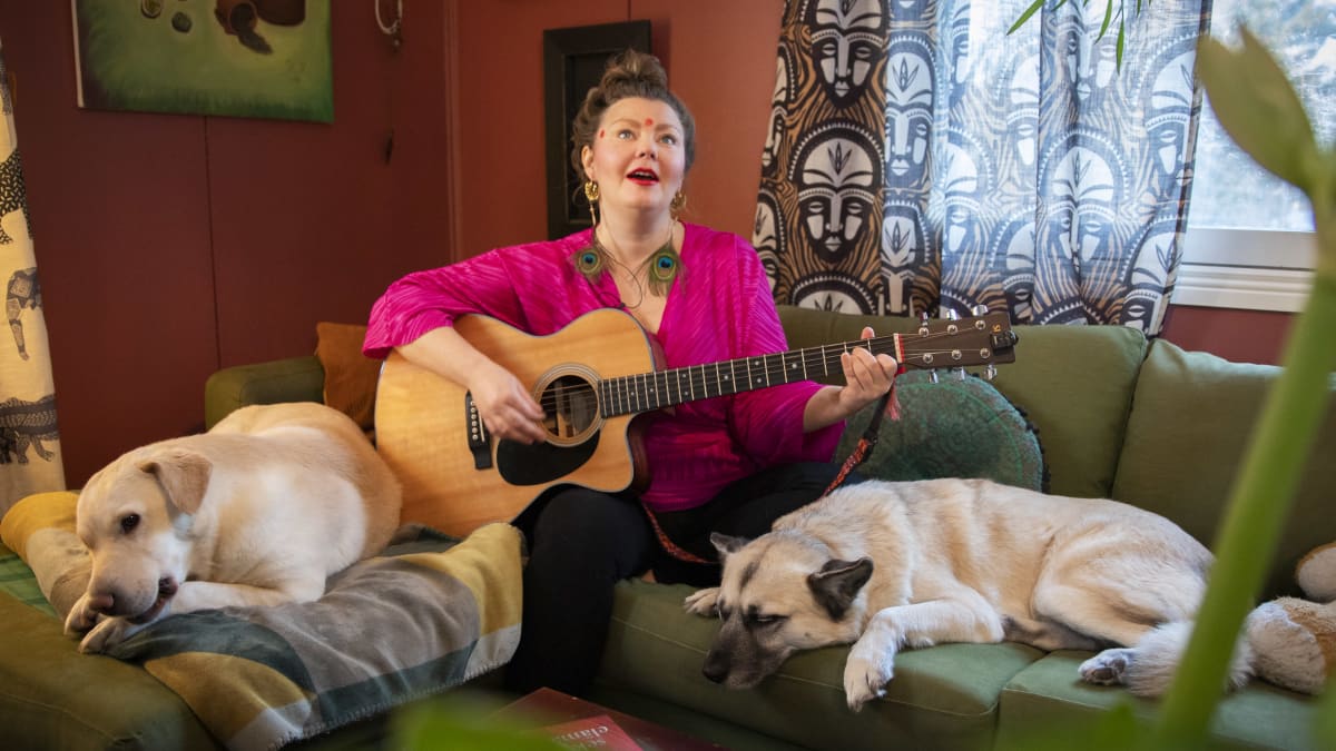 Heiduska soittaa kitaraa ja laulaa kotisohvallaan koirat vierellään.