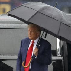 Yhdysvaltain entinen presidentti Donald Trump pitää mustaa sateenvarjoa päänsä päällä, takanaan musta tila-auto.