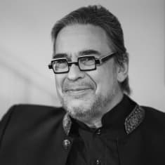 En svartvit bild av Jyrki Kasvi som lutar mot en vägg samtidigt som han tittar in i kameran och ler. Han är klädd i en svart skjorta och svart kavaj.