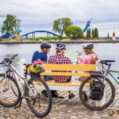 Kolme ihmistä istuu puiston penkillä järvimaisemassa ja tutkii karttaa. Penkin ympärillä on kolme polkupyörää.