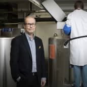 Professori Heikki Hyöty seisoo tutkimuslaboratoriossa. Vieressä työntekijä seisoo säiliön luona.