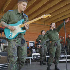 Kuvassa maastopukuihin pukeutunut bändi soittamassa. Etualalla sinistä kitaraa soittava mies ja mikrofonia pitelevä nainen. 