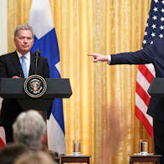 Two men in dark suits standing behind podiums with Finnish and US flags behind them, the larger man on the right pointing to the man on the left..