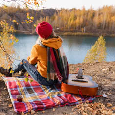 Henkilö istuu kalliolla ruudullisella huovalla kitara vieressään ja katsoo edessään näkyvää järveä ja syksyistä metsää.