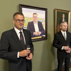 Kaksi miestä seisoo muotokuvamaalauksen vierellä lasit kädessään.