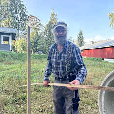 Ruutupaitainen harmaapartainen mies seisoo maaseudun pihapiirissa pitkä puukeppi kädessä. 