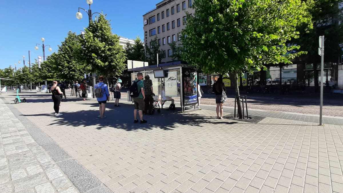 Ihmisiä seisoo kaupungin kadulla bussipysäkin vieressä. Osa odottaa lehmuksen varjossa. Taustalla näkyy turkoosi sähköpotkulauta.