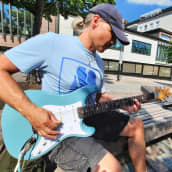 Lippalakkipäinen mies soittaa helteisenä kesäpäivänä kitaraa Kokkolan kävelykadulla.