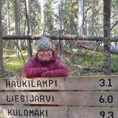 Leena Löf nojaa puiseen reittiopastekylttiin taustalla Seitsemisen kansallispuiston kuusimetsää ja kaatuneita puita
