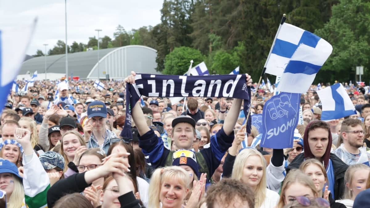 Yleisömerta hymyilemässä ja taputtamassa. Yleisön keskellä mies nostamassa huivia, jossa lukee "Finland".