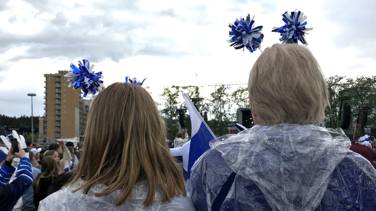 Takaapäin otettu kuva kahdesta tytöstä seisomassa sadeviitat päällä sateessa. Tytöillä päässä Suomi-teemaiset "tuntosarvet".