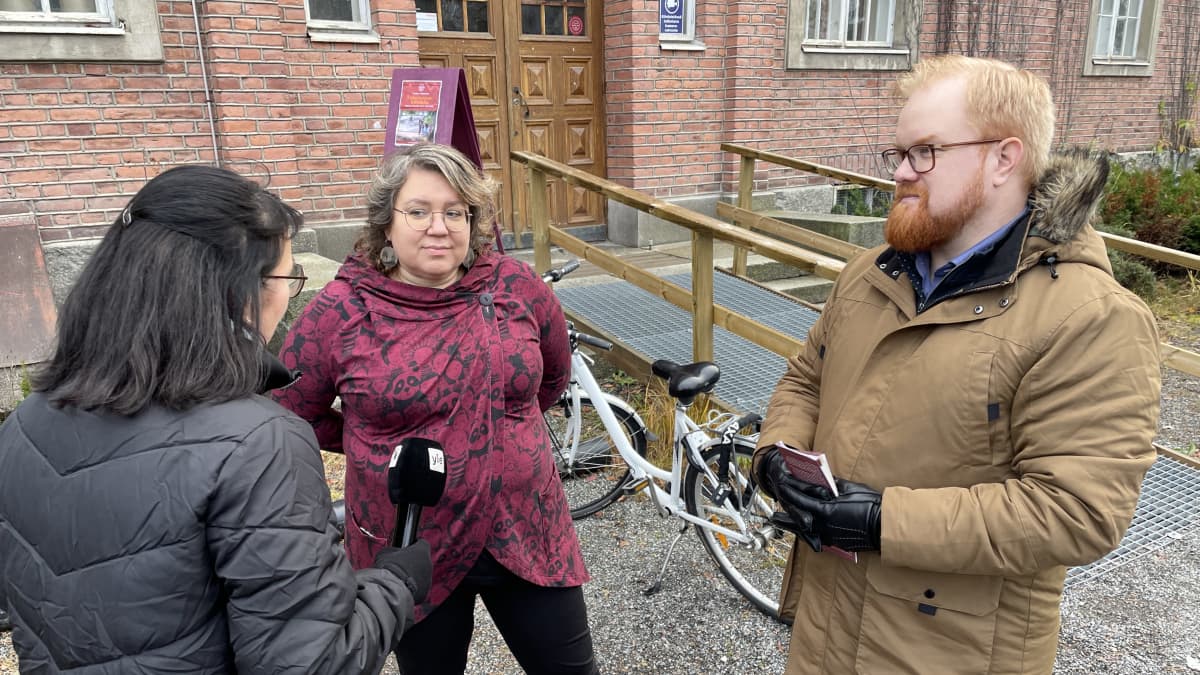 Punaisiin pukeutunut nainen ja ruskeaan pukeutunut mies tummatukkaisen toimittajan haastattelussa punaisen tiilitalon soraisella pihalla.