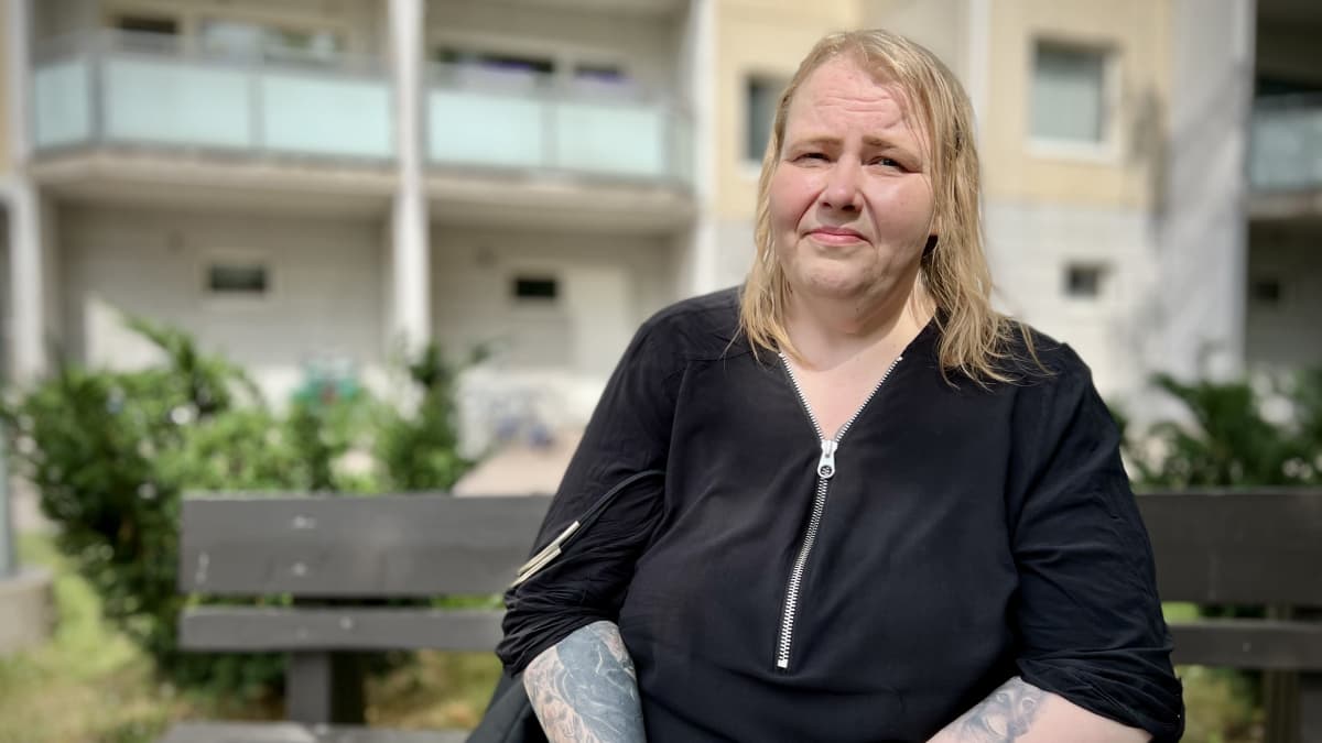 Marianne Mäkinen istuu kerrostalon pihassa ruskealla penkillä.