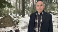 Pappi seisoo lumisen muistolehdon edessä. Taustalla kuusia ja hautakynttilälyhtyjä.