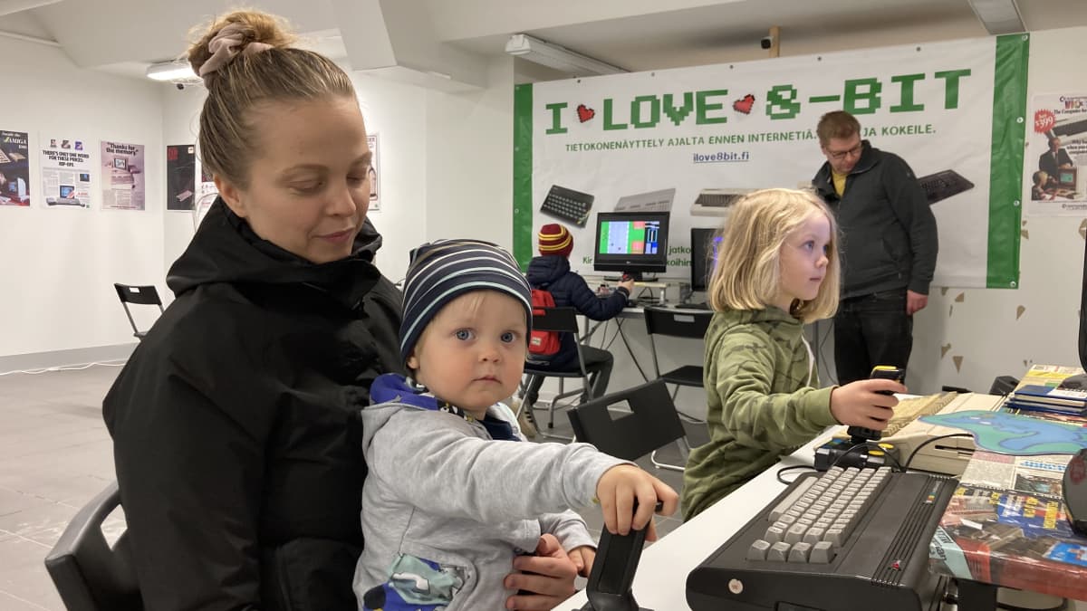 Lapsi istuu naisen sylissä ja pelaa tietokonepeliä joystickillä.