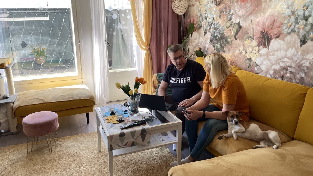Janne ja Yana Suutari keskustelevat kotinsa sohvalla istuen. Vieressä makaa perheen koira. 