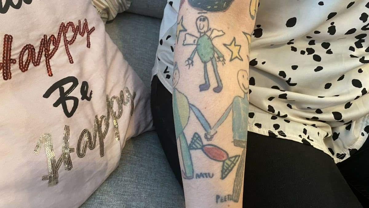 Lasten piirustuksia tatuoituna äidin käteen, kaksoset pitämässä toisiaan kädestä ja yläpuolella leijailee toinen pojan kuolemansa jälkeen enkelinä, kuvassa näkyy myös lakritsikuorrutteinen donitsi.