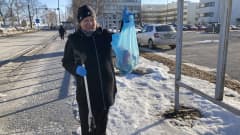 Nainen nostaa sinistä roskapussia, johon on kerääjän avulla noukkinut roskia kevättalviselta kadulta kaupungilla.